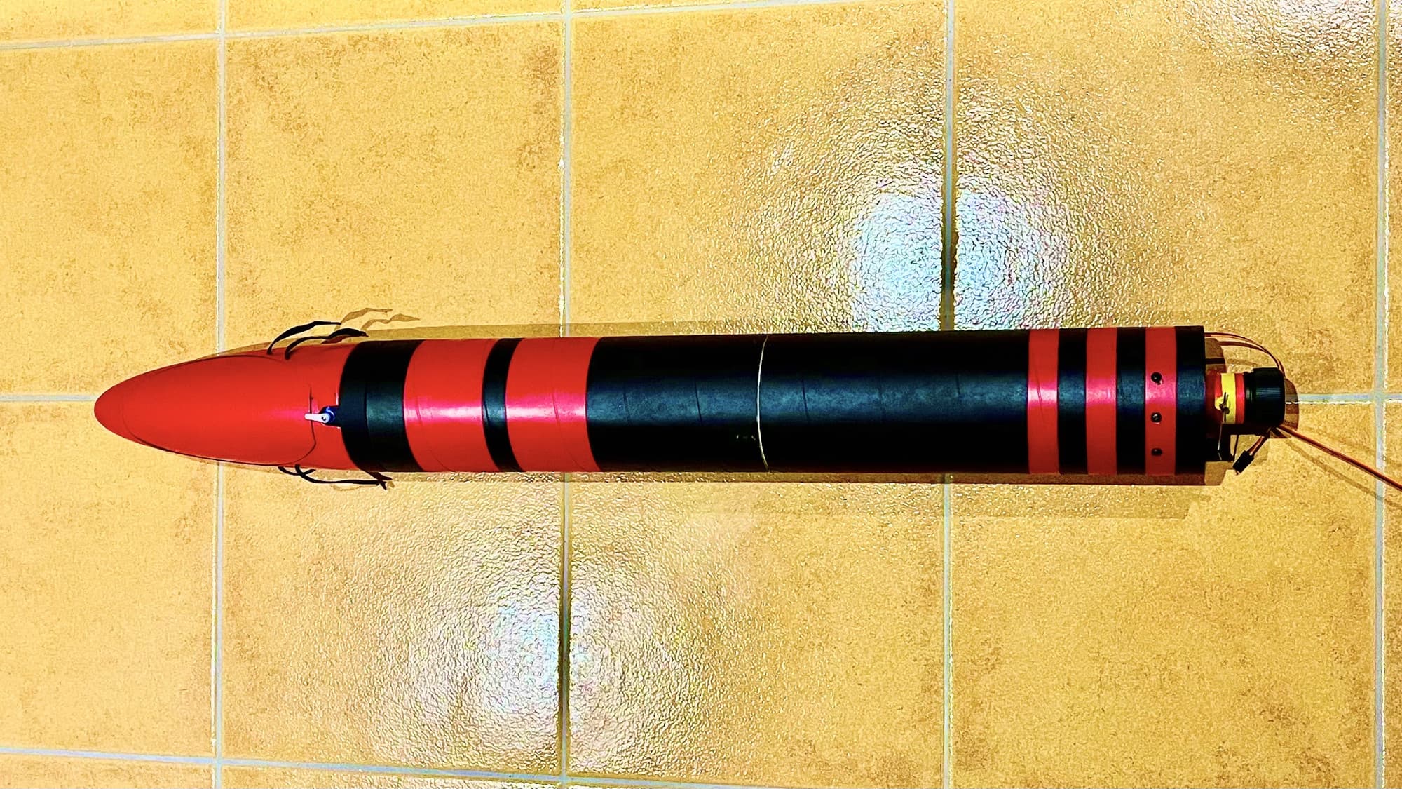 The finished finless model rocket lying on a tiled floor, red nose cone and black-and-red banded body, nose to tail